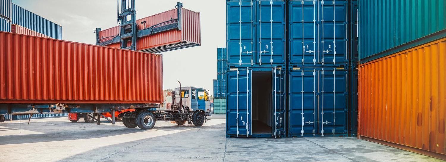 Ein Gabelstapler hebt einen roten Versandcontainer auf einem belebten Containerhof an, in dessen Nähe blaue und orangefarbene Container gestapelt sind.