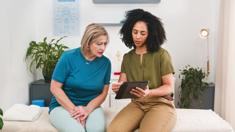 Eine Physiotherapeutin sitzt neben ihrer Patientin auf einer Liege und zeigt ihr etwas auf einem Tablet