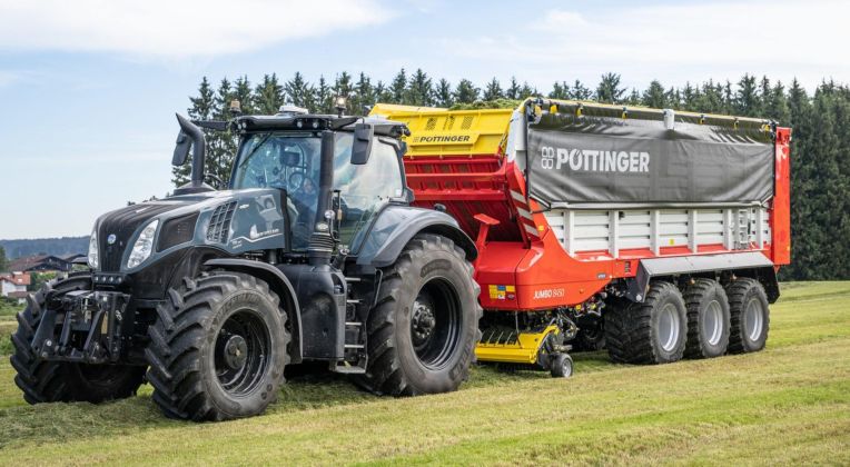 Moderner schwarzer Traktor mit großem PÖTTINGER JUMBO 8450 Ladewagen auf einem frisch gemähten Feld vor einem Waldhintergrund.