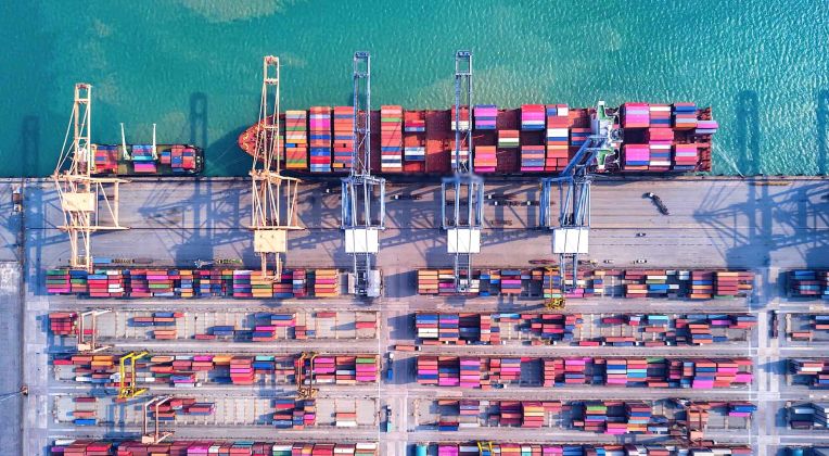 Luftaufnahme eines Containerhafens mit gestapelten Containern, großen Kränen und einem Frachtschiff im türkisfarbenen Wasser.