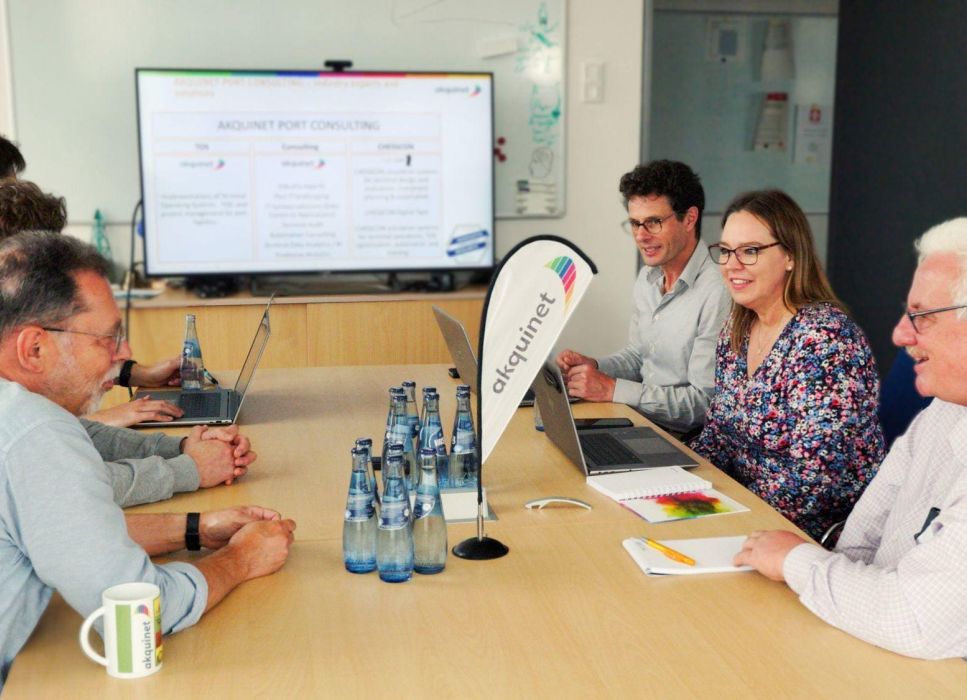 Ein Team von Fachleuten sitzt in einem Konferenzraum, diskutiert über Hafenlogistik und betrachtet eine Präsentation auf einem Bildschirm.